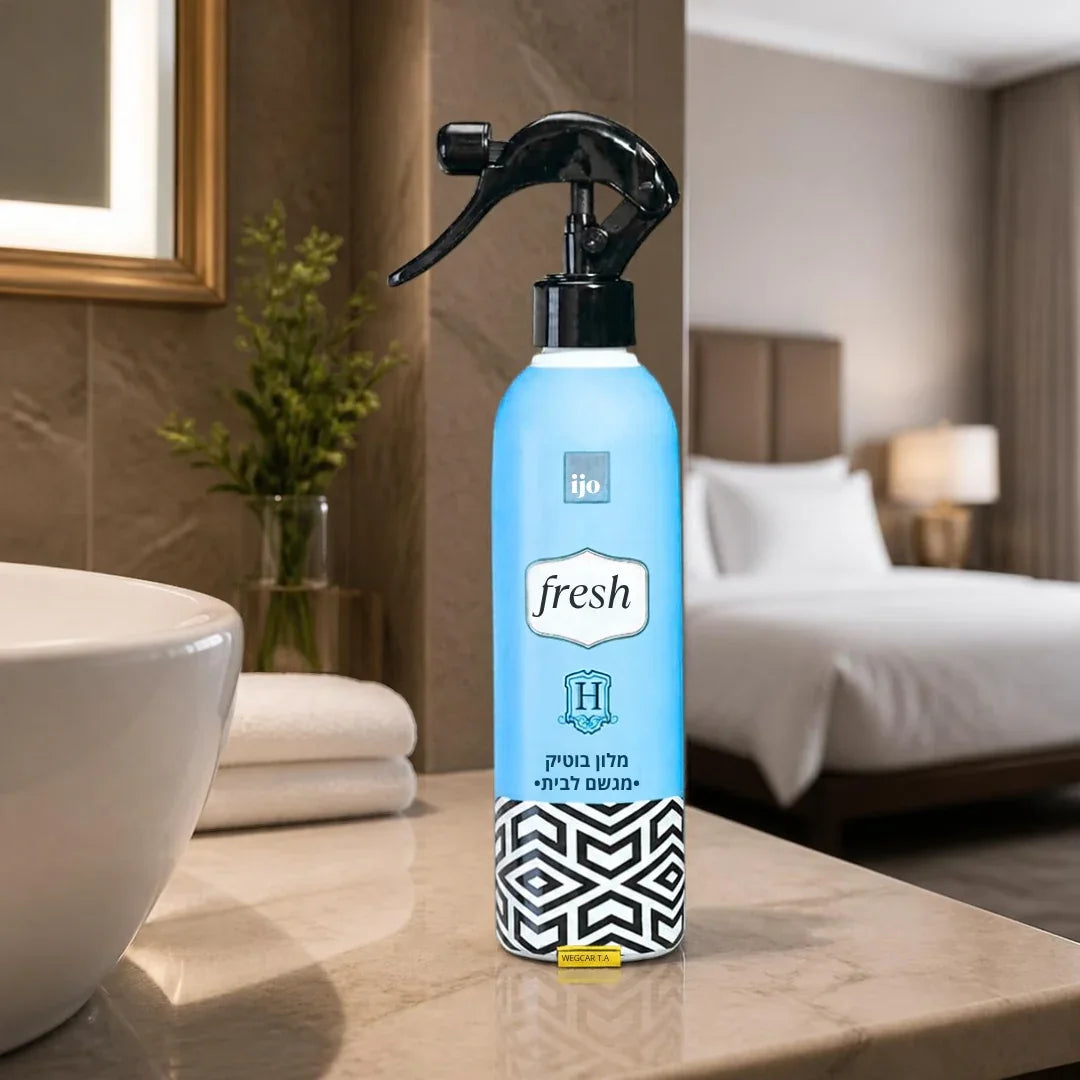 A light blue spray bottle of ijo Fresh room spray with a black trigger sprayer, standing on a marble bathroom counter with a blurred hotel room background.