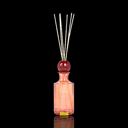 Pink glass fragrance diffuser bottle with wooden reeds and a red decorative topper.