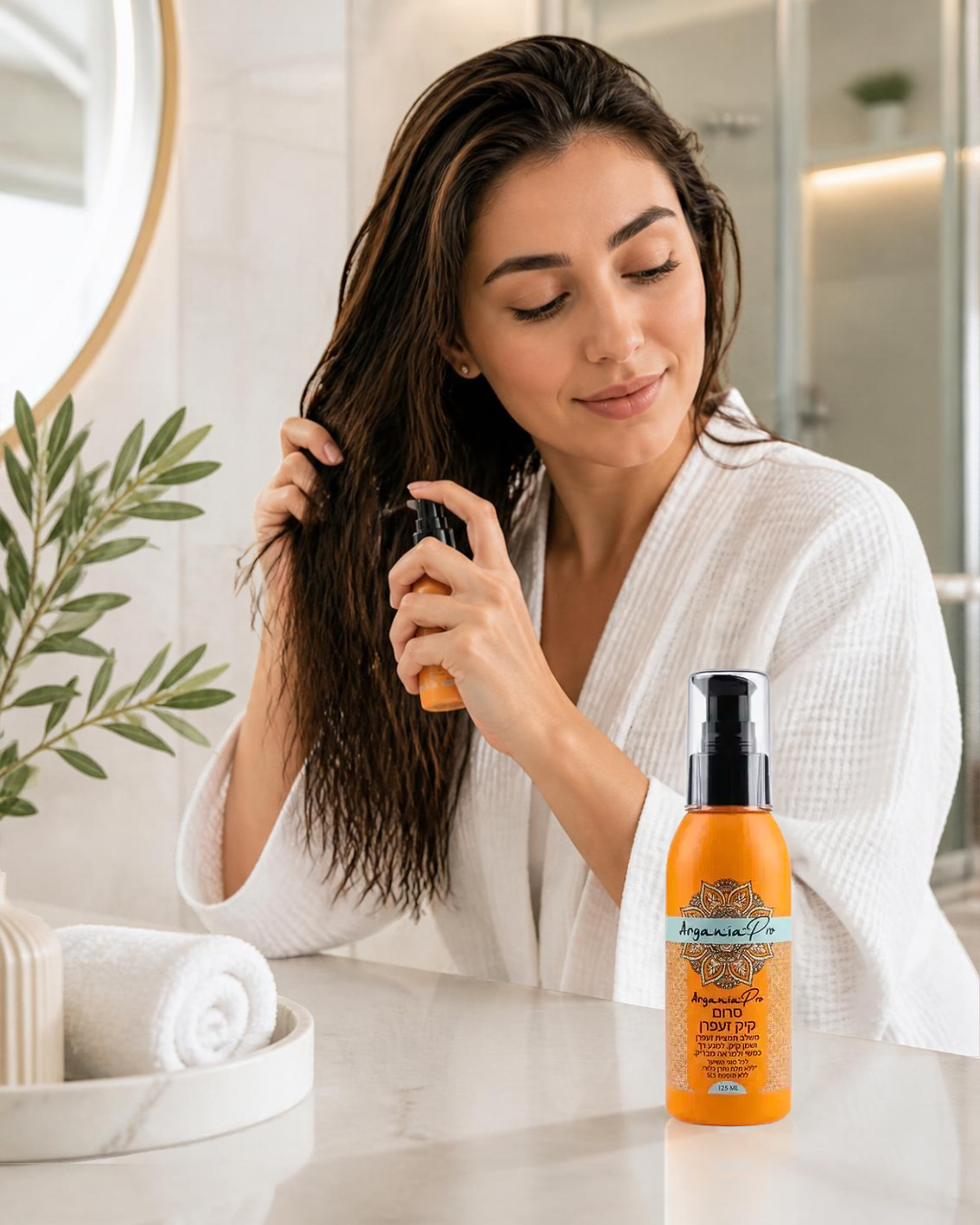 Woman in a white robe applying Castor Saffron Hair Serum to damp hair, with the orange bottle on a marble counter