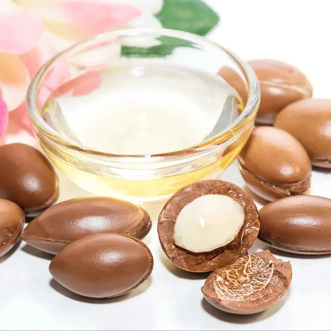 Argan kernels around a small glass bowl of golden argan oil on a white surface