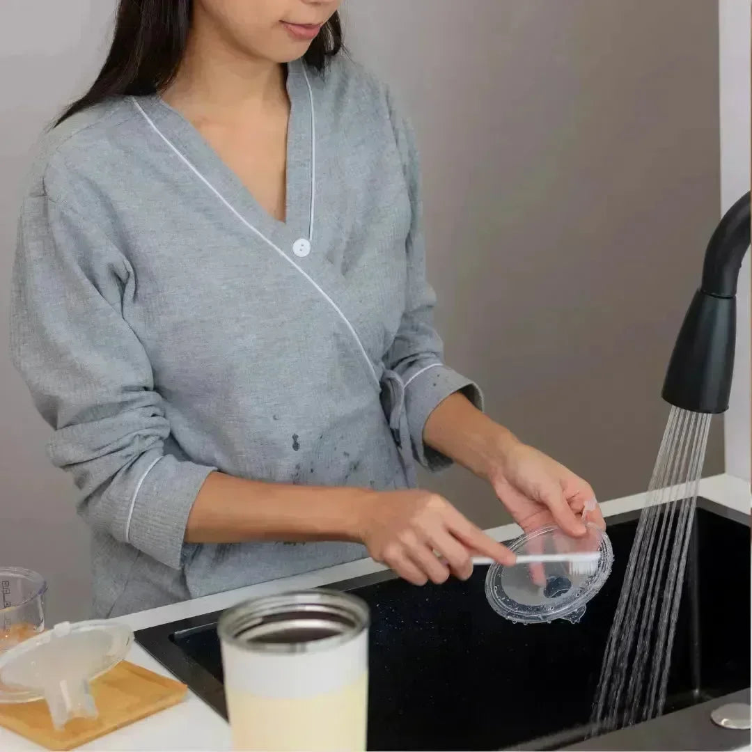 Woman rinsing a clear baby bottle part with a small brush under running tap water at a sink