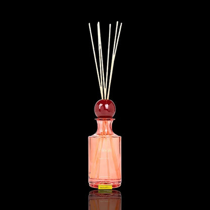 Pink glass fragrance diffuser bottle with wooden reeds and a red decorative topper.