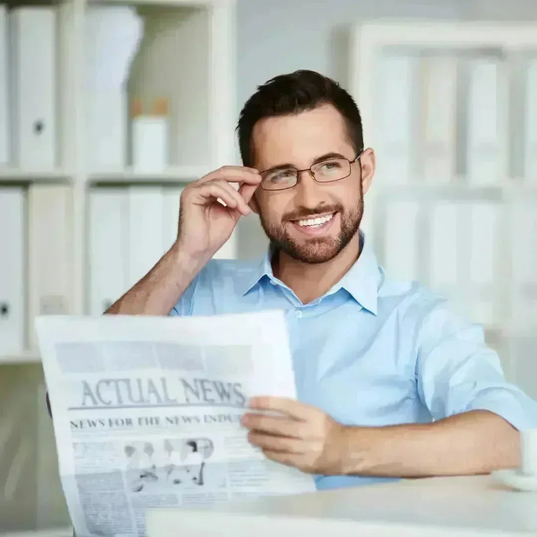 Smiling man in a light blue shirt and glasses holding a newspaper at a desk